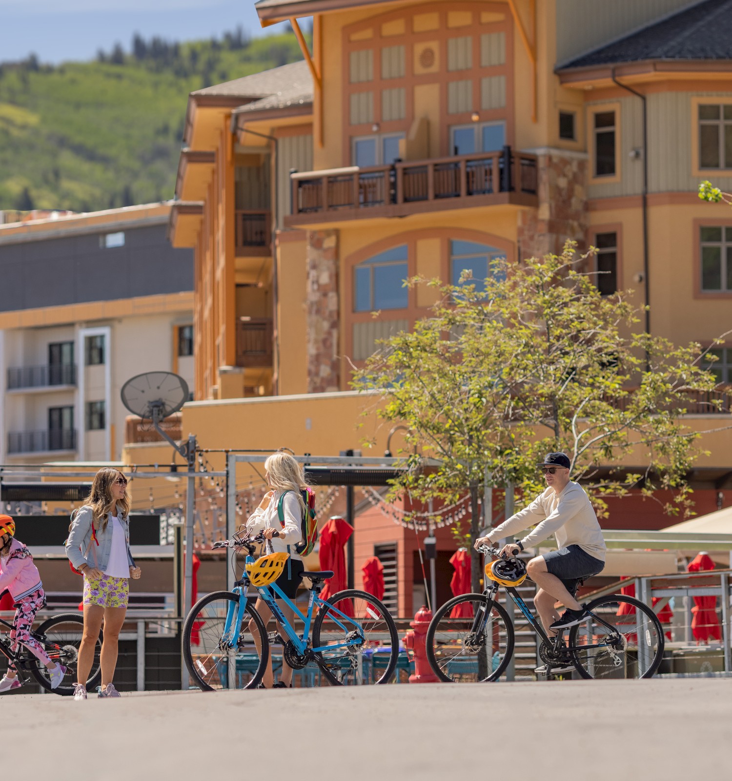 People are bicycling on a sunny street near buildings with hills in the background.