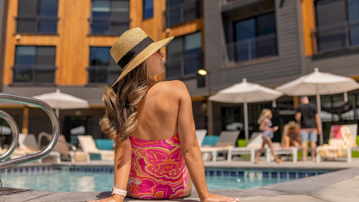 A person in a hat and bright swimsuit sits by a pool at a modern apartment complex with people and umbrellas in the background.
