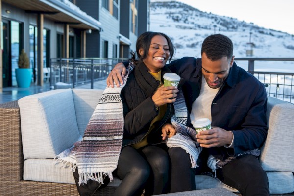 A smiling couple sits on a patio sofa, wrapped in a blanket, holding drinks. Snowy mountains are visible in the background.
