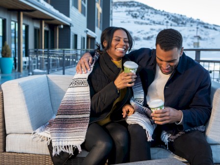 A couple sits on an outdoor sofa, wrapped in a blanket, holding cups with snowy mountains in the background, sharing a joyful moment.