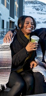 A couple is sitting outside, laughing and holding coffee cups, with a snowy mountain in the background.
